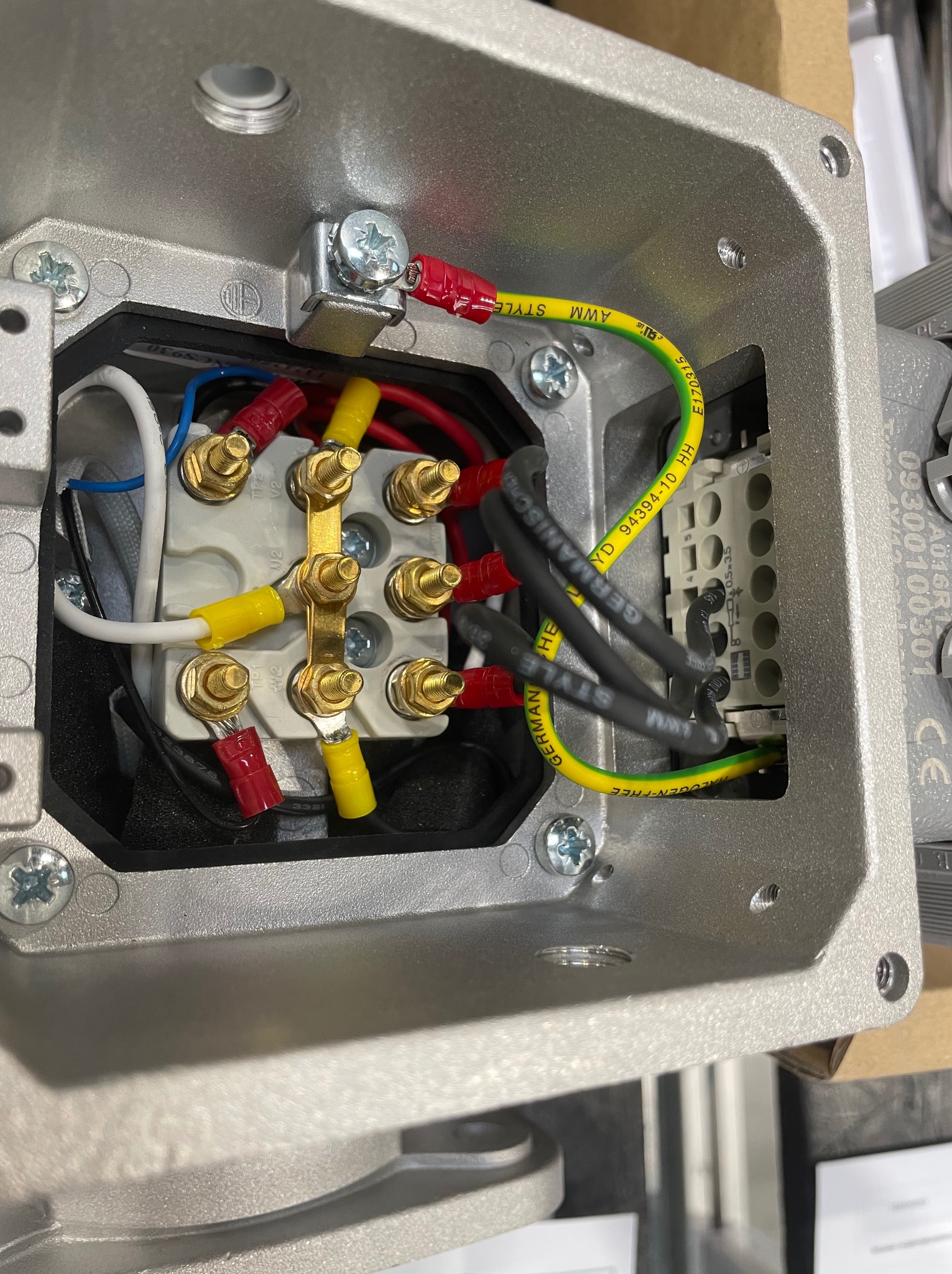 Wnętrze metalowej puszki przyłączeniowej z widocznymi przewodami elektrycznymi zakończonymi konektorami w kolorach czerwonym, żółtym i niebieskim, podłączonymi do listwy zaciskowej. Widoczny...