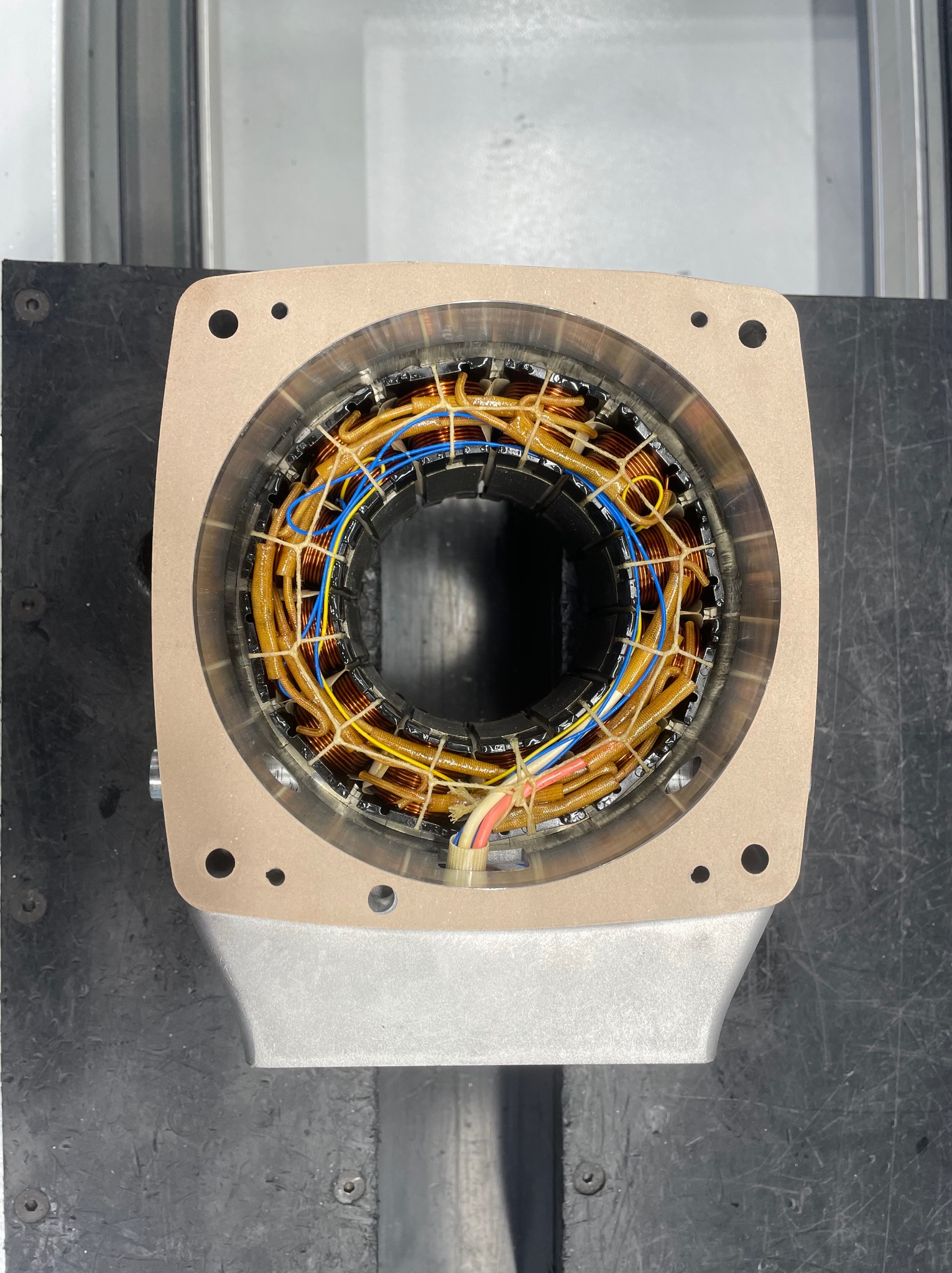 Wnętrze silnika elektrycznego z widocznymi uzwojeniami miedzianymi w izolacji, niebieskimi i żółtymi przewodami oraz elementami mocującymi, widok z góry na warsztatowym stole.