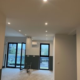 Wykończenia wnętrz @ Designe - Wnętrze mieszkania w trakcie prac wykończeniowych: widoczne narzędzia malarskie, surowa podłoga, pomalowane ściany do połowy wysokości na ciemny kolor, okna balkonowe z widokiem na zieleń.