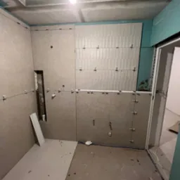 Łazienka w trakcie remontu: częściowo ułożone białe płytki na ścianie, widoczne krzyżyki dystansowe, ściany obłożone płytami kartonowo-gipsowymi, sufit z elementami gładzi, widoczne otwory...