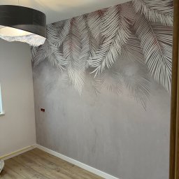 Wykończenia wnętrz @ Designe - Ściana w pokoju z szarym tynkiem dekoracyjnym i motywem białych liści palmowych, widoczny fragment drewnianej podłogi i czarna lampa z abażurem.