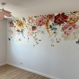 Wykończenia wnętrz @ Designe - Biała ściana w pokoju z naklejonym motywem kwiatowym, drewniana podłoga, widoczny fragment lampy w kształcie kwiatu i gniazdko elektryczne.