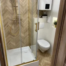 Nowoczesna łazienka z kabiną prysznicową ze złotymi elementami, toaletą i bojlerem na ścianie, wykończona płytkami imitującymi drewno ułożonymi w jodełkę.