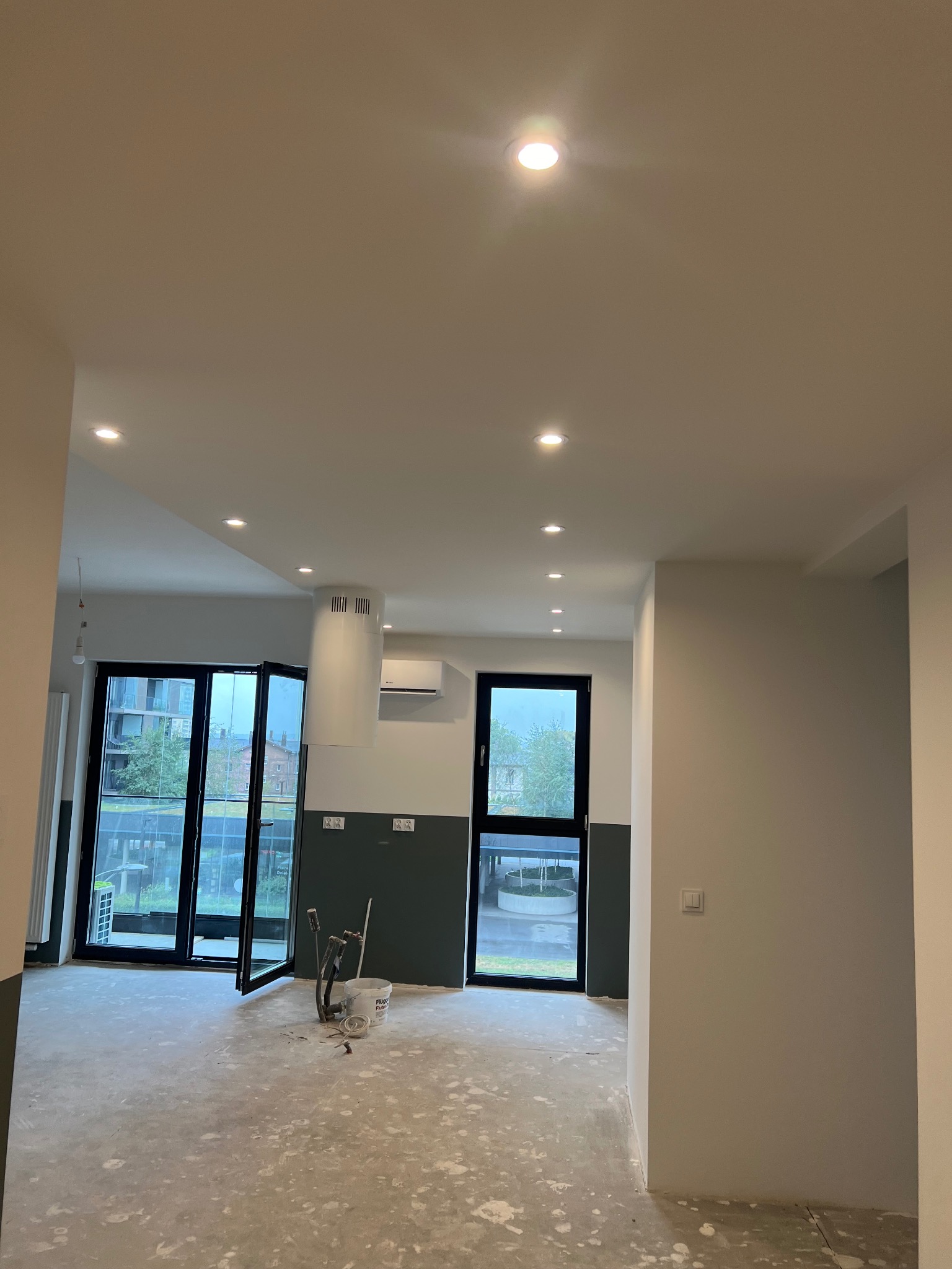 Wnętrze mieszkania w trakcie prac wykończeniowych: widoczne narzędzia malarskie, surowa podłoga, pomalowane ściany do połowy wysokości na ciemny kolor, okna balkonowe z widokiem na zieleń.