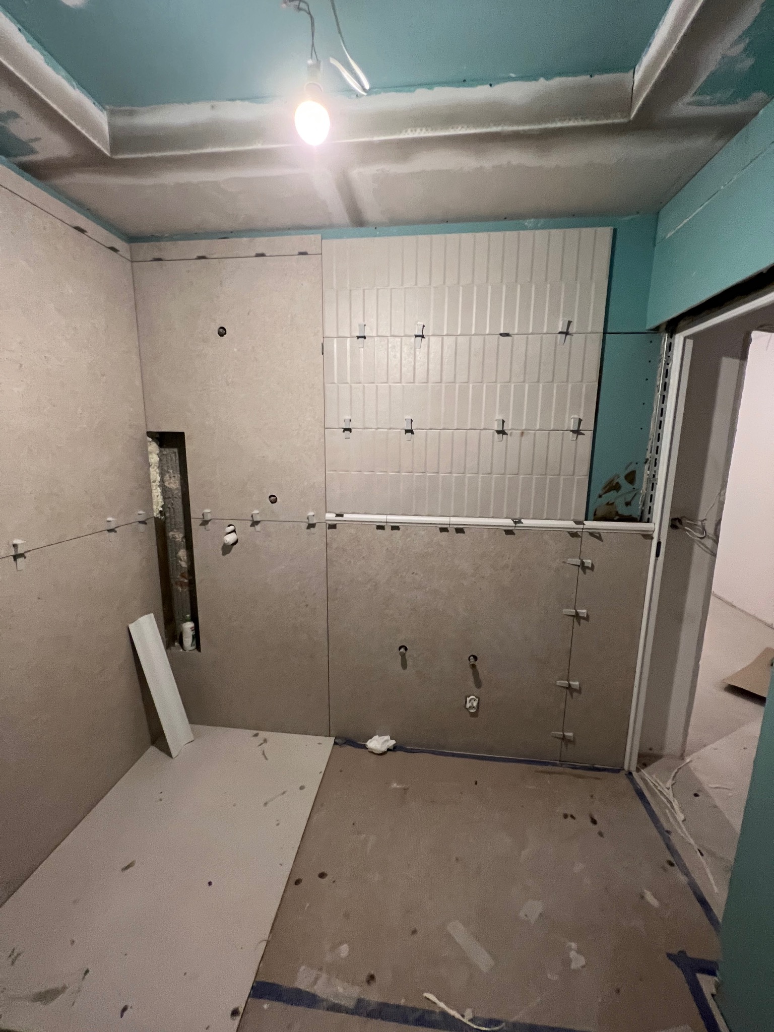 Łazienka w trakcie remontu: częściowo ułożone białe płytki na ścianie, widoczne krzyżyki dystansowe, ściany obłożone płytami kartonowo-gipsowymi, sufit z elementami gładzi, widoczne otwory...