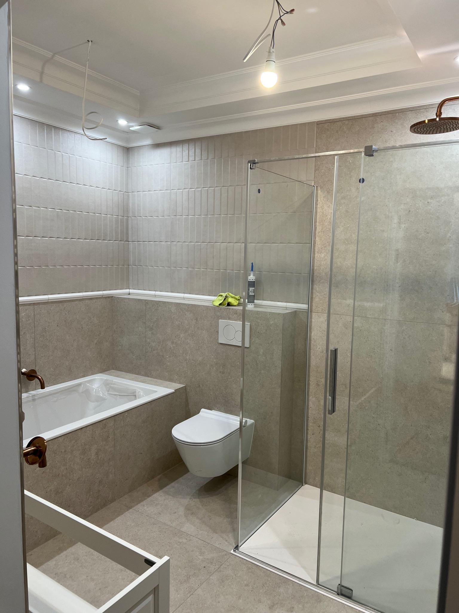 Nowoczesna łazienka w trakcie wykańczania: beżowe płytki, kabina prysznicowa ze szklanymi drzwiami, wanna z obudową, podwieszana toaleta, armatura w kolorze miedzi, widoczne przewody elektryczne...