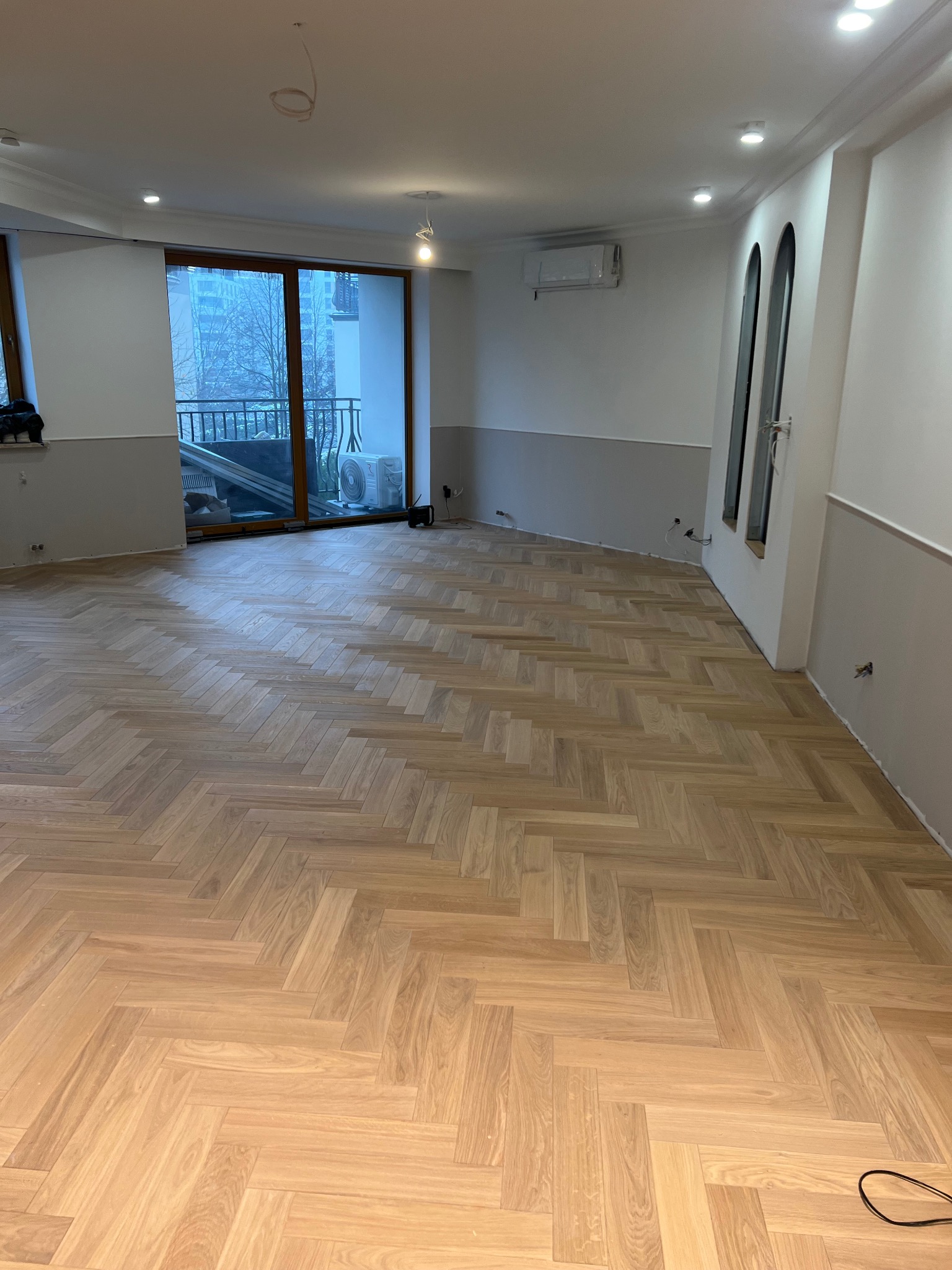 Jasny parkiet ułożony we wzór jodełki w przestronnym pokoju z oknem balkonowym i szarymi panelami ściennymi.