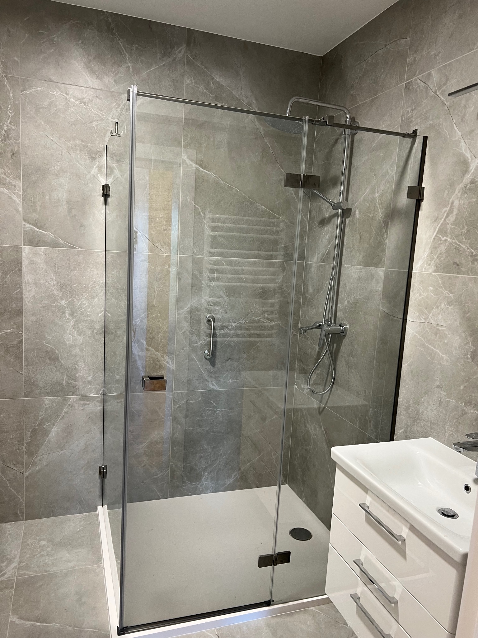 Szklana kabina prysznicowa z deszczownicą i baterią, zamontowana w łazience wyłożonej szarymi płytkami imitującymi marmur, z umywalką na szafce w rogu.