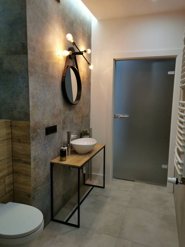 Nowoczesna łazienka z umywalką nablatową na drewnianym blacie z czarną metalową konstrukcją, oświetlona dekoracyjną lampą z żarówkami, szara płytka na ścianie i podłodze, matowe szklane drzwi.