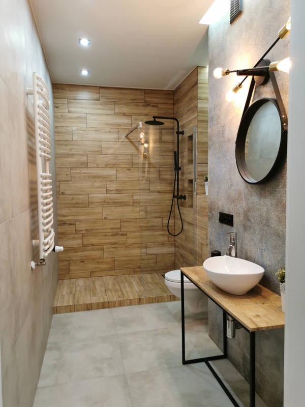 Nowoczesna łazienka z drewnopodobnymi płytkami, kabiną prysznicową typu walk-in, umywalką nablatową na drewnianym blacie z metalowym stelażem oraz okrągłym lustrem z dekoracyjnym oświetleniem.