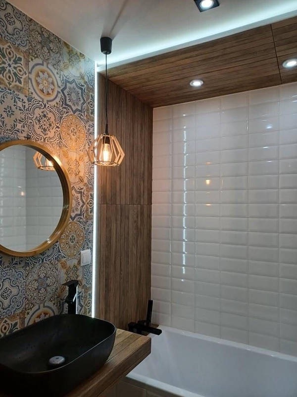 Wykończona łazienka z dekoracyjnymi płytkami ściennymi, okrągłym lustrem w złotej ramie, czarną umywalką na drewnianym blacie i podświetlanym sufitem z drewnianą boazerią.