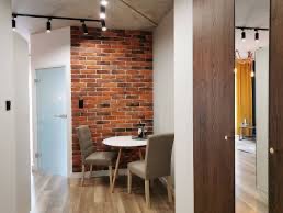 Wnętrze mieszkania z ceglaną ścianą, okrągłym białym stolikiem i dwoma krzesłami na drewnianej podłodze. Widoczne oświetlenie sufitowe i fragment lustra odbijającego zasłony.