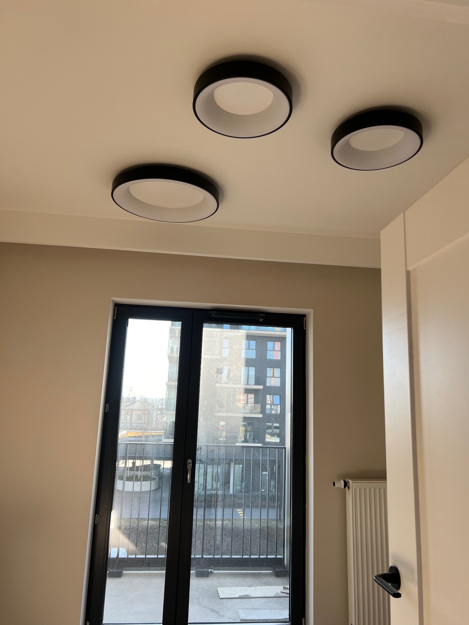 Wnętrze pokoju z trzema okrągłymi lampami sufitowymi z czarną obwódką, widok na balkon przez czarne drzwi balkonowe, beżowe ściany, białe drzwi po prawej stronie.