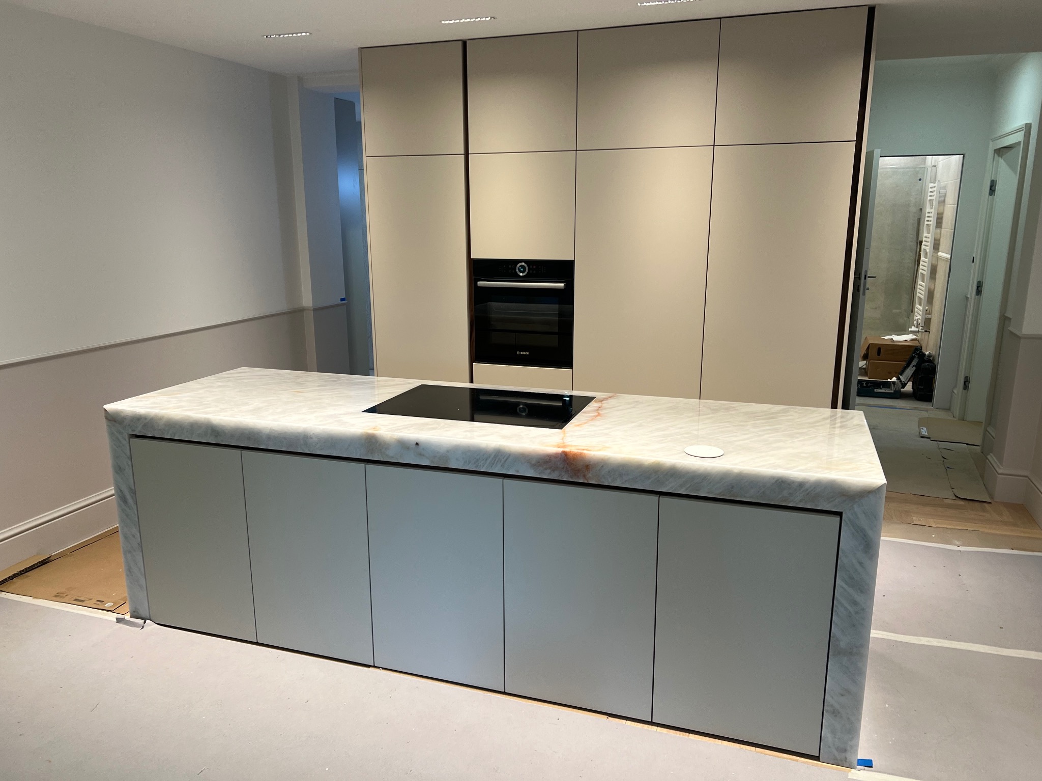 Wnętrze kuchni w trakcie remontu: zabudowa kuchenna z beżowymi szafkami i marmurowym blatem wyspy, widoczny piekarnik w zabudowie i płyta indukcyjna, podłoga zabezpieczona folią malarską.