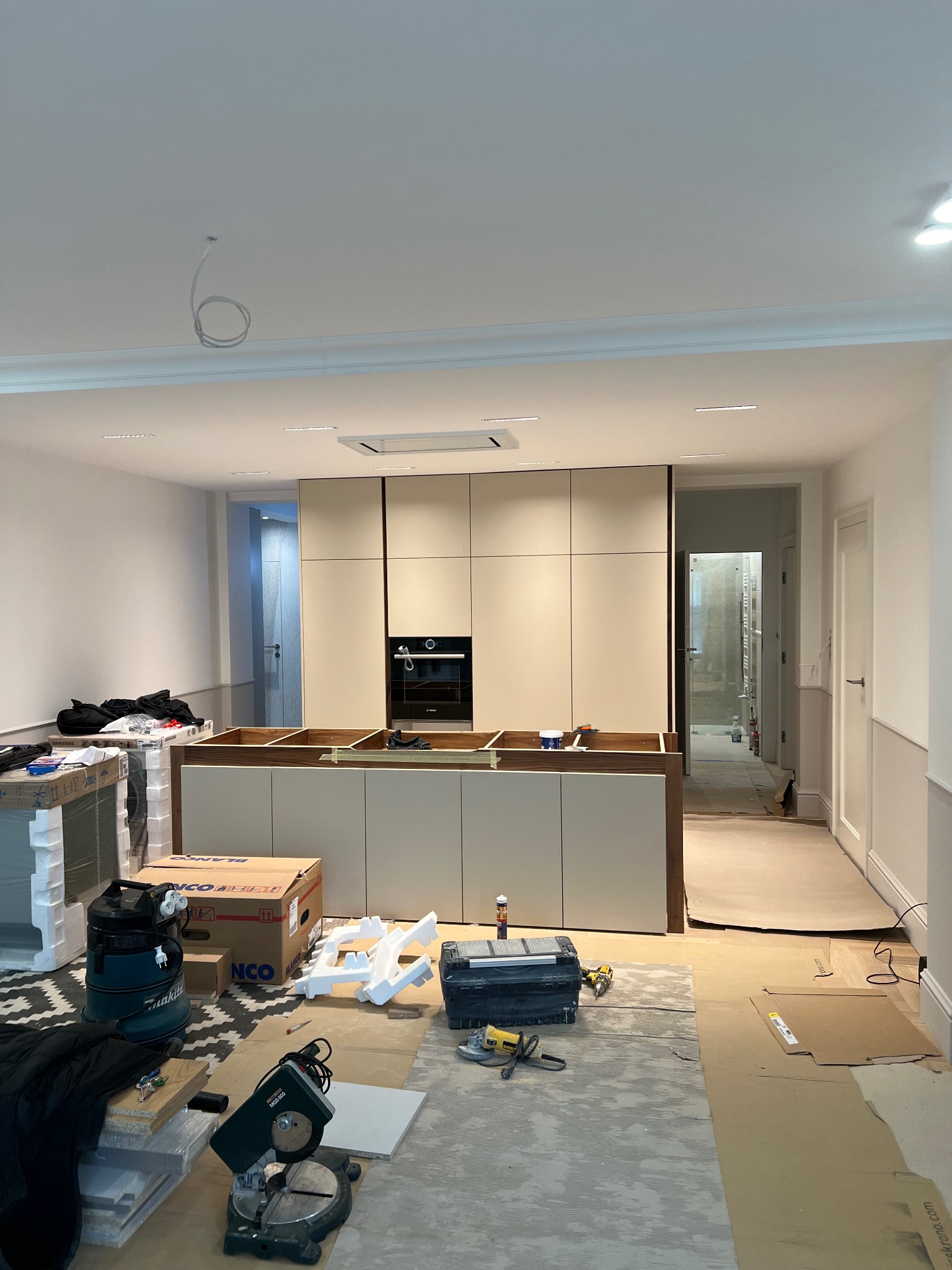 Wnętrze w trakcie prac wykończeniowych: zabudowa kuchenna z zamontowanym piekarnikiem, widoczne narzędzia, kartony i elementy montażowe na podłodze zabezpieczonej kartonem.