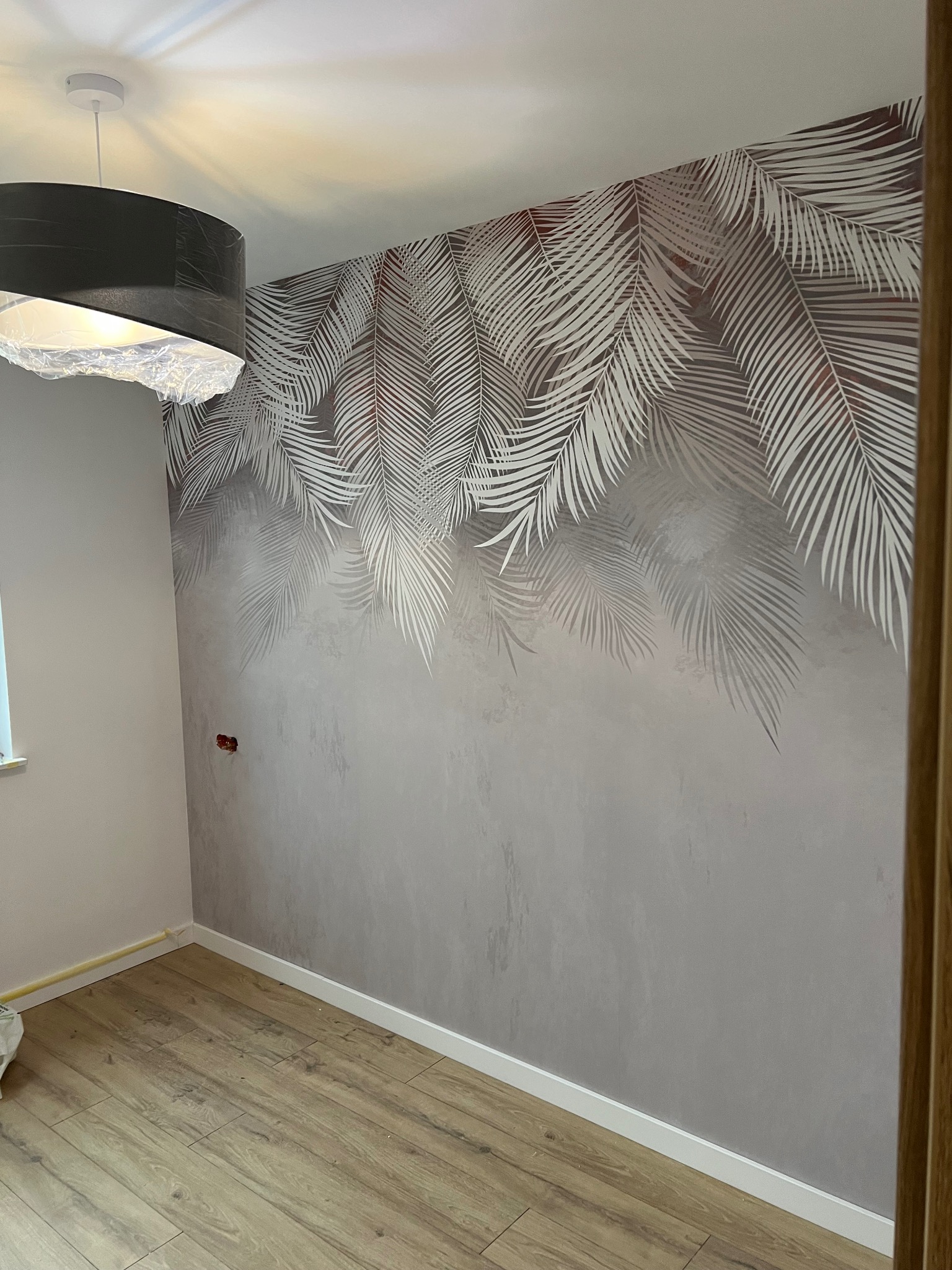 Ściana w pokoju z szarym tynkiem dekoracyjnym i motywem białych liści palmowych, widoczny fragment drewnianej podłogi i czarna lampa z abażurem.