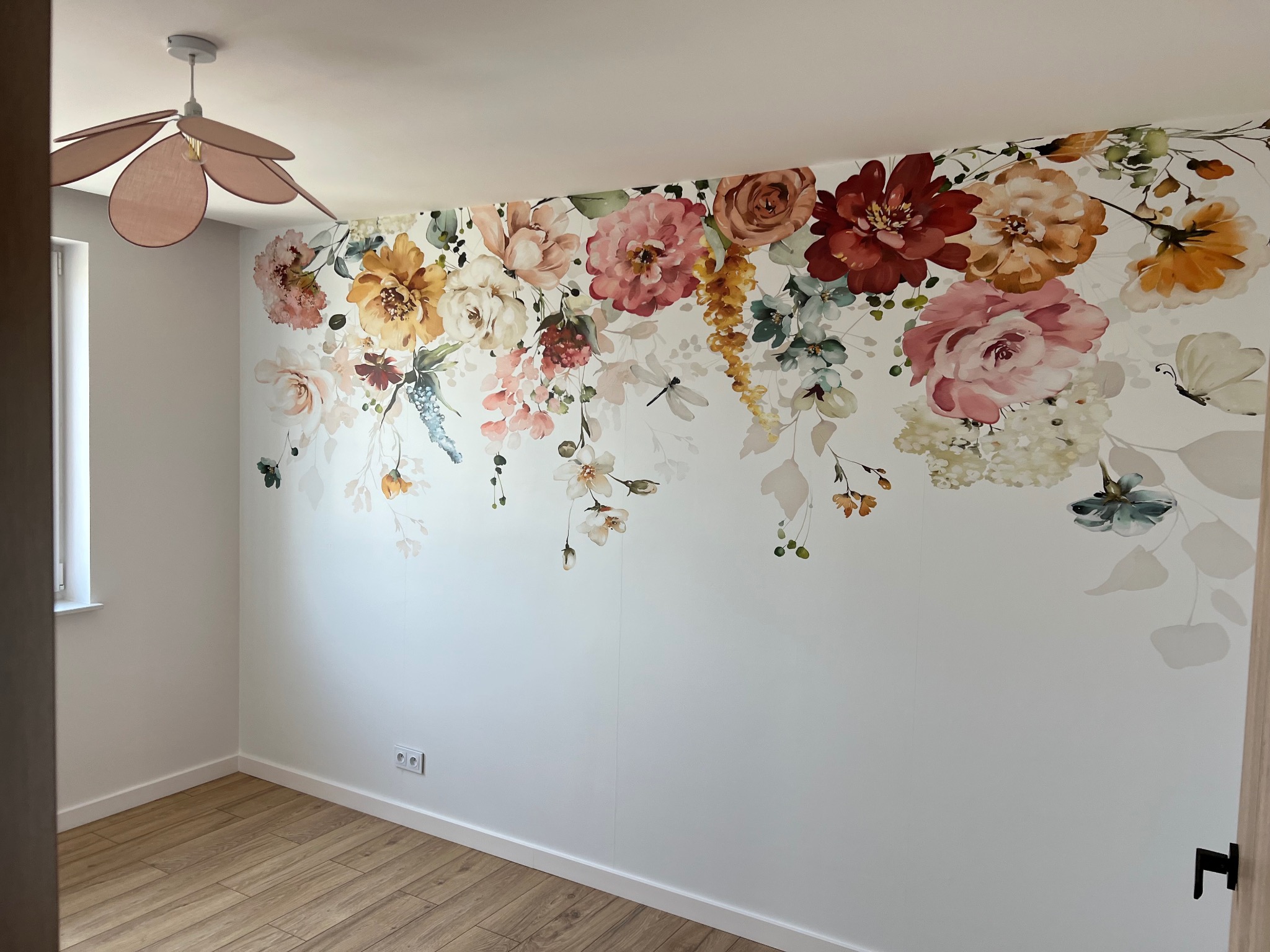 Biała ściana w pokoju z naklejonym motywem kwiatowym, drewniana podłoga, widoczny fragment lampy w kształcie kwiatu i gniazdko elektryczne.