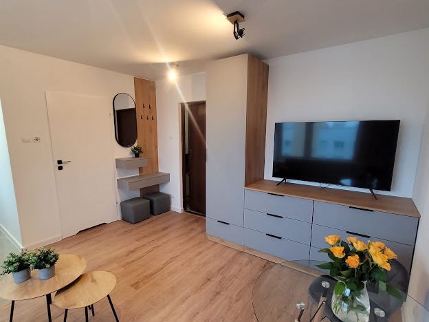 Jasne wnętrze mieszkania po remoncie: drewniana podłoga, szare meble, telewizor na ścianie, dekoracje roślinne i bukiet żółtych róż na szklanym stole.