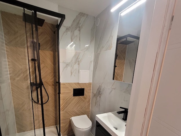 Nowoczesna łazienka z kabiną prysznicową typu walk-in z czarną armaturą, umywalką nablatową i toaletą w zabudowie, wykończona płytkami imitującymi marmur i drewno.