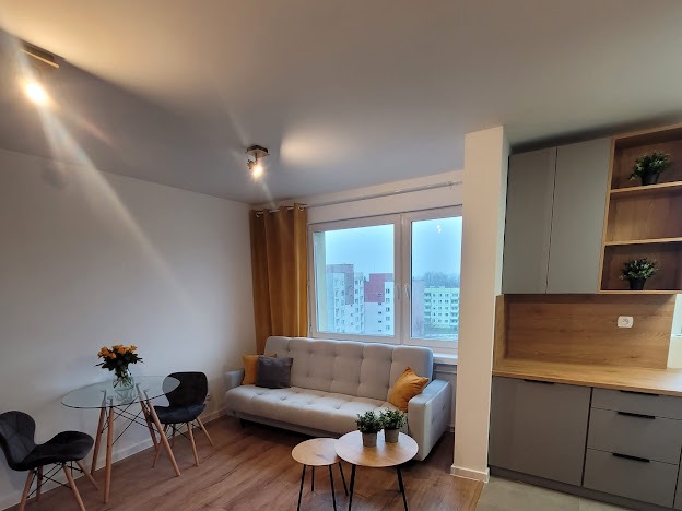 Nowoczesne wnętrze mieszkania z drewnianą podłogą, szarą sofą z poduszkami, szklanym stołem z kwiatami i aneksem kuchennym w odcieniach szarości i drewna.