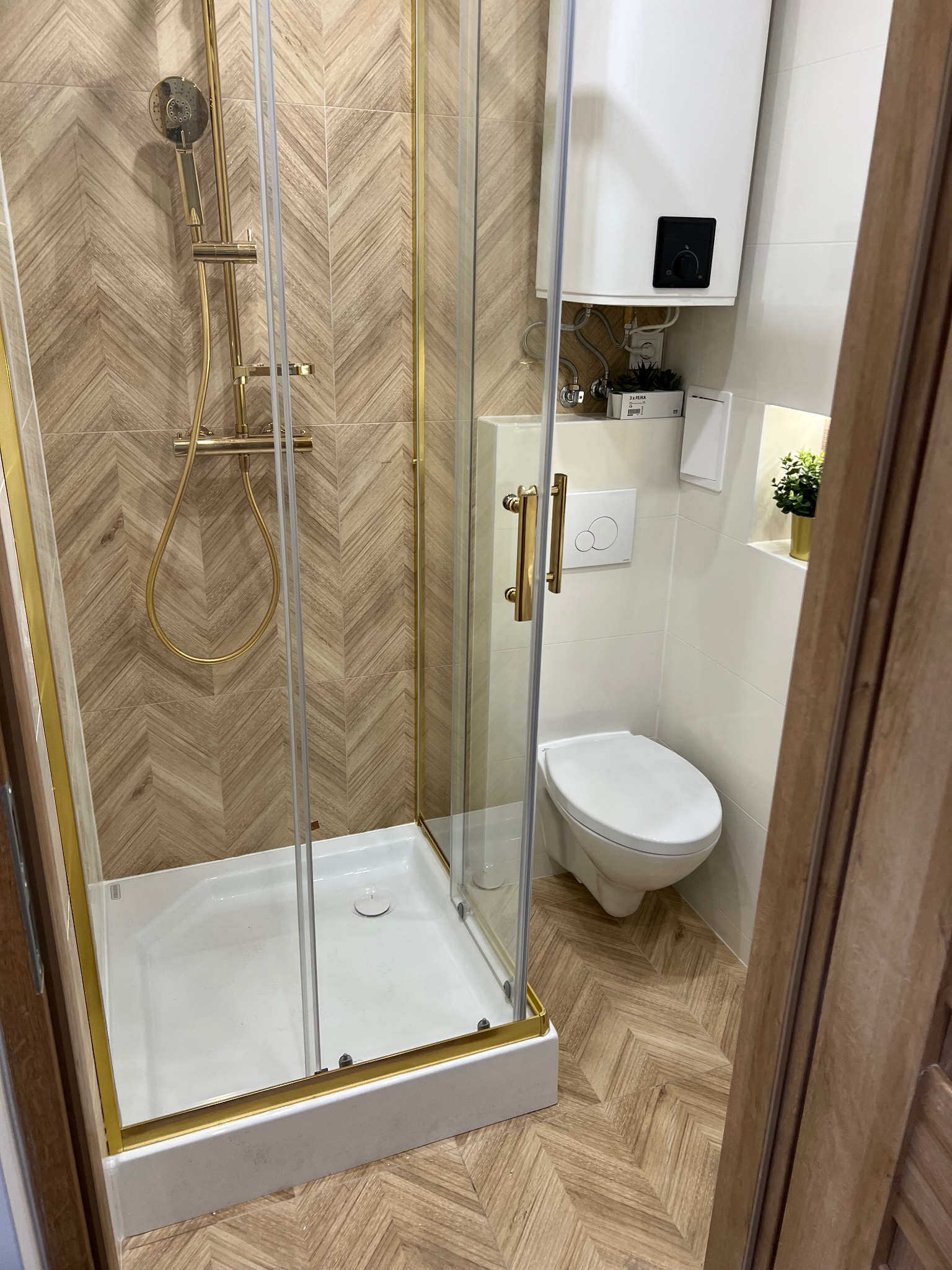 Nowoczesna łazienka z kabiną prysznicową ze złotymi elementami, toaletą i bojlerem na ścianie, wykończona płytkami imitującymi drewno ułożonymi w jodełkę.