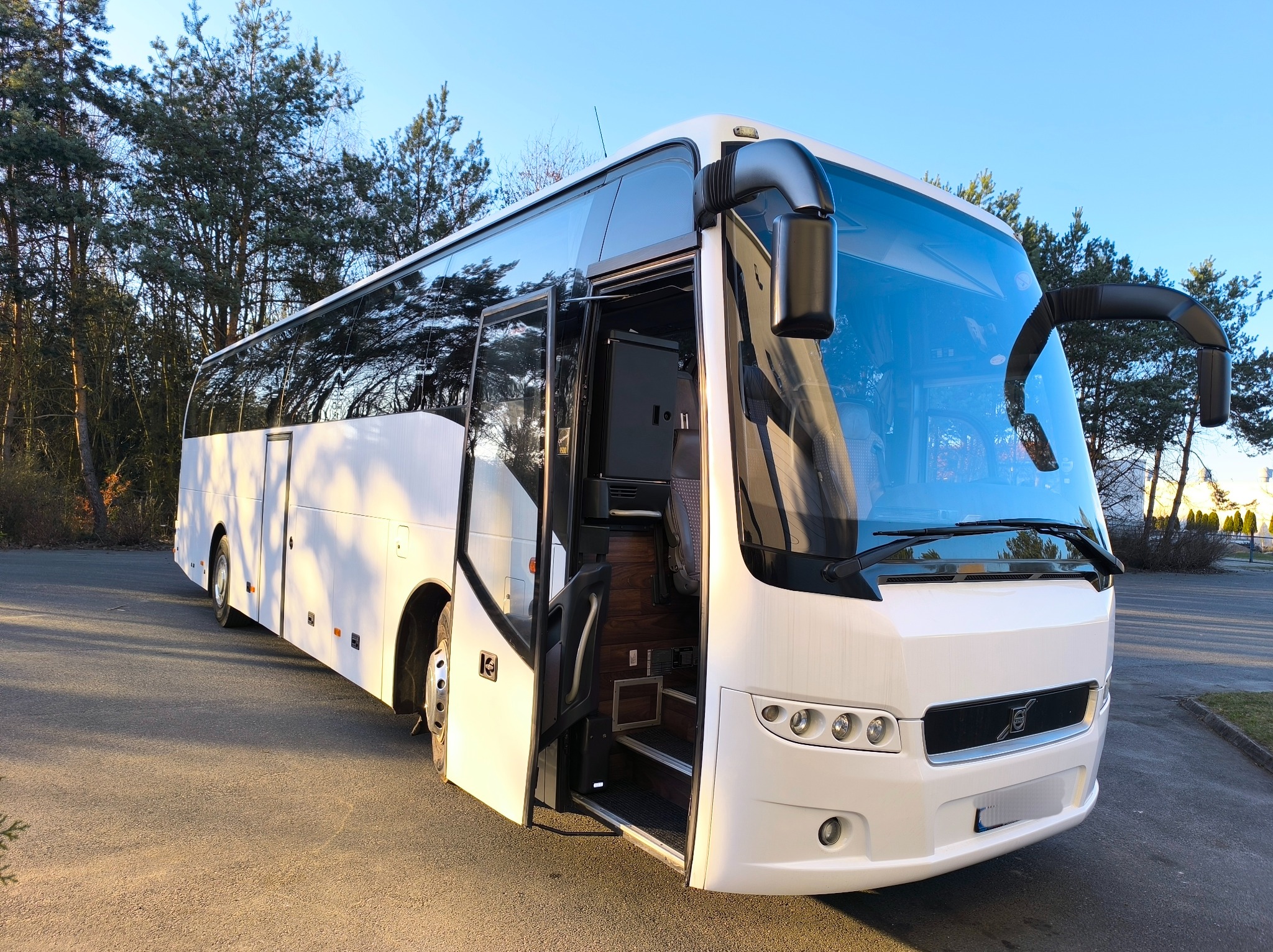 Nowoczesny, biały autokar turystyczny z otwartymi drzwiami, zaparkowany na parkingu z widocznym lasem w tle.