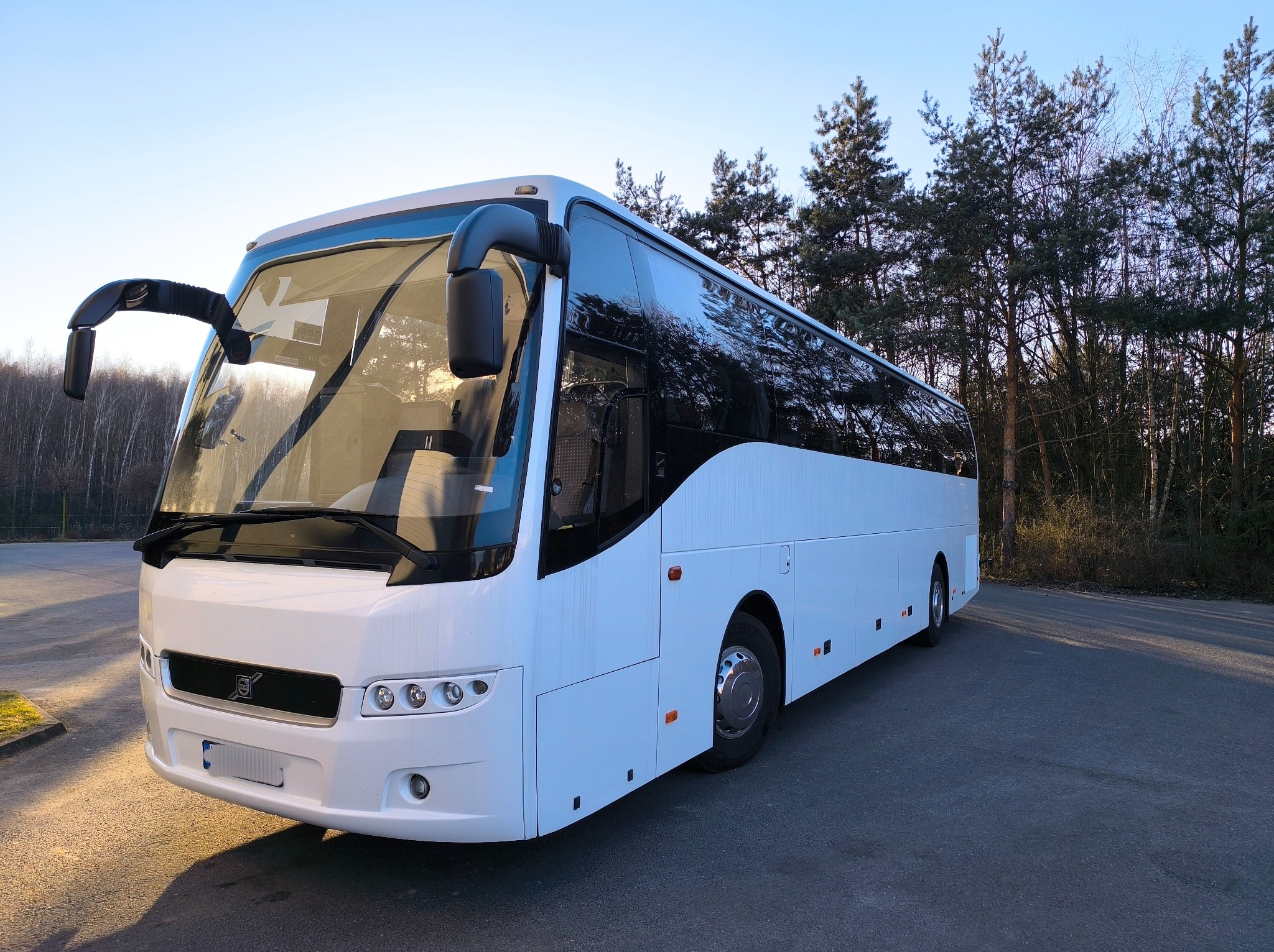 Biały autokar turystyczny zaparkowany na asfaltowym placu, z lasem w tle, widok z przodu i boku.