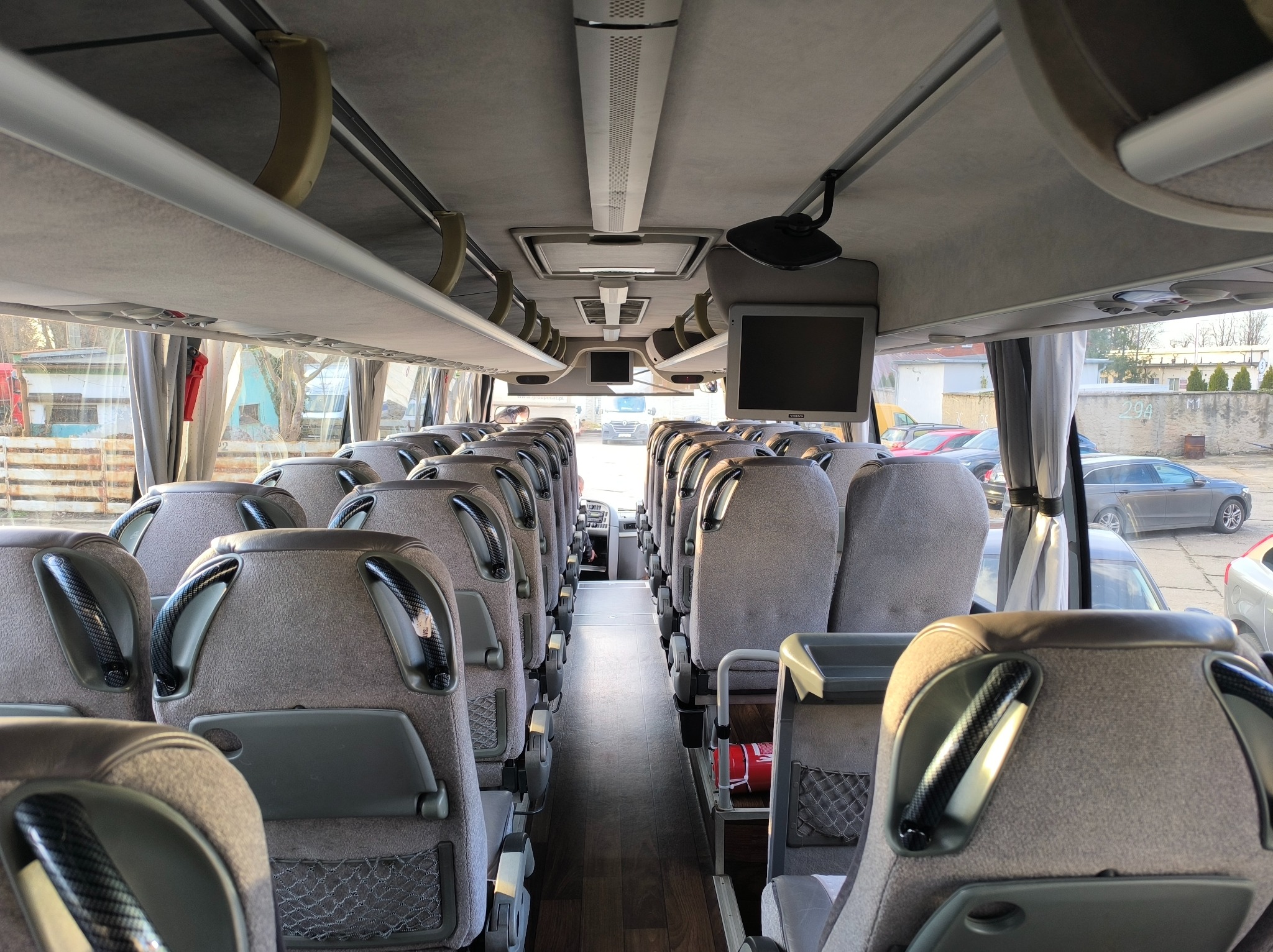 Wnętrze autobusu turystycznego z rzędami szarych foteli z uchwytami w karbonowym wzorze, widok z tyłu pojazdu w kierunku przedniej szyby i monitorów podwieszonych pod sufitem.