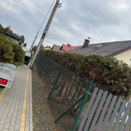 Przyczepa z zielonymi gałęziami zaparkowana na chodniku obok żywopłotu i szarego ogrodzenia panelowego, widok na domy pod pochmurnym niebem.