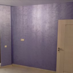 Serhii Ryzhenko - Ściana w pokoju pomalowana dekoracyjną farbą w odcieniach fioletu i szarości, widoczne gniazdka elektryczne i fragment białych drzwi z kluczem w zamku.