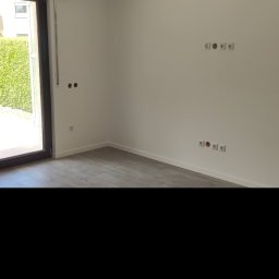 Serhii Ryzhenko - Pusty, świeżo wyremontowany pokój z szarą podłogą imitującą drewno, białymi ścianami z gniazdkami elektrycznymi i otwartym balkonem z widokiem na zielony żywopłot.