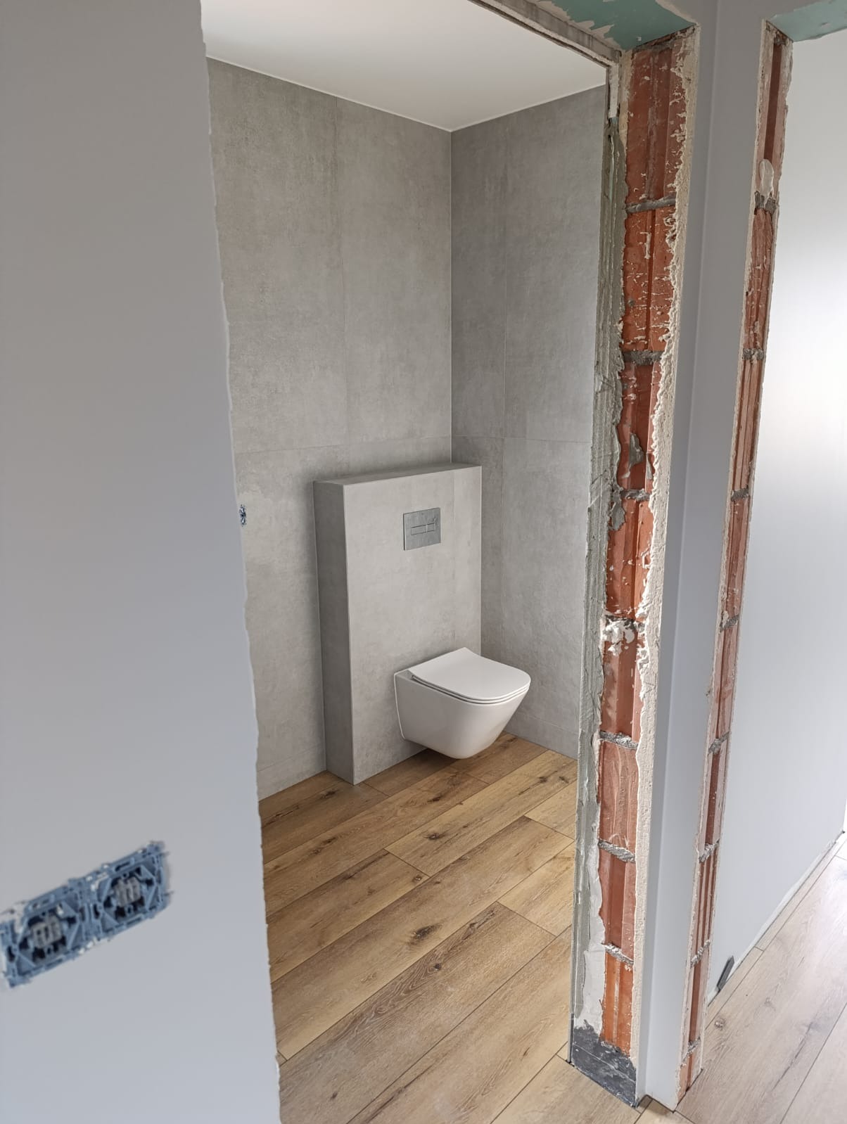 Wisząca toaleta z podtynkową spłuczką w szarej obudowie, szare płytki na ścianach, drewnopodobne panele na podłodze, widoczne niedokończone ościeże drzwi z cegieł i zaprawy.