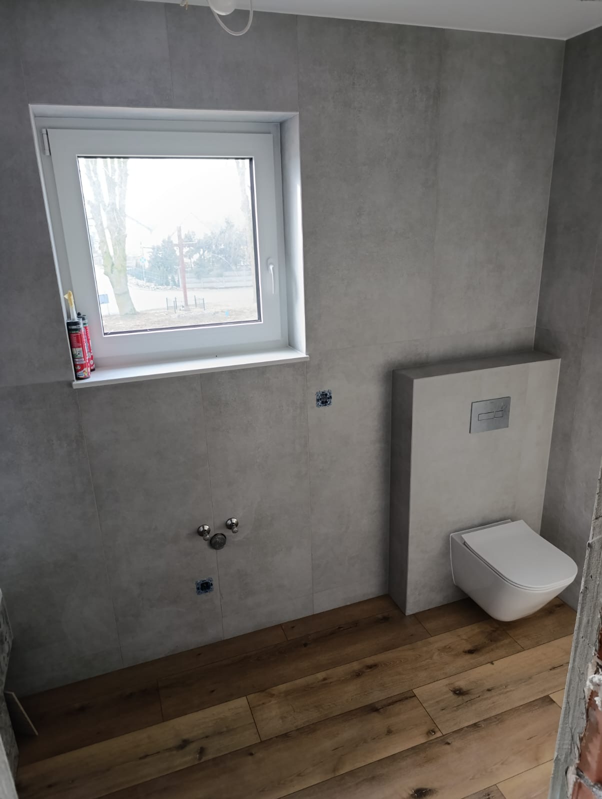 Nowoczesna łazienka w trakcie wykańczania: szare płytki wielkoformatowe na ścianach, drewnopodobna podłoga, wisząca miska WC z zabudowanym stelażem, okno z widokiem na zewnątrz, puszki elektryczne...