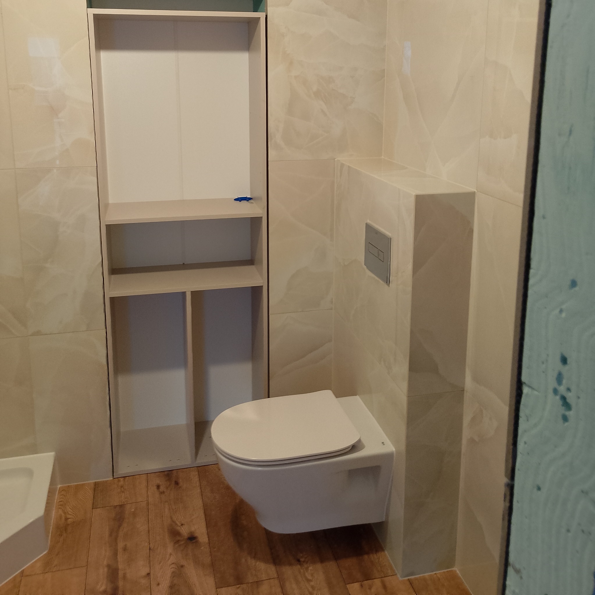 Nowoczesna łazienka w trakcie wykańczania: podwieszana toaleta na tle beżowych płytek imitujących marmur, drewnopodobna podłoga i zabudowa z półkami. Widoczny fragment zabezpieczonej ściany.