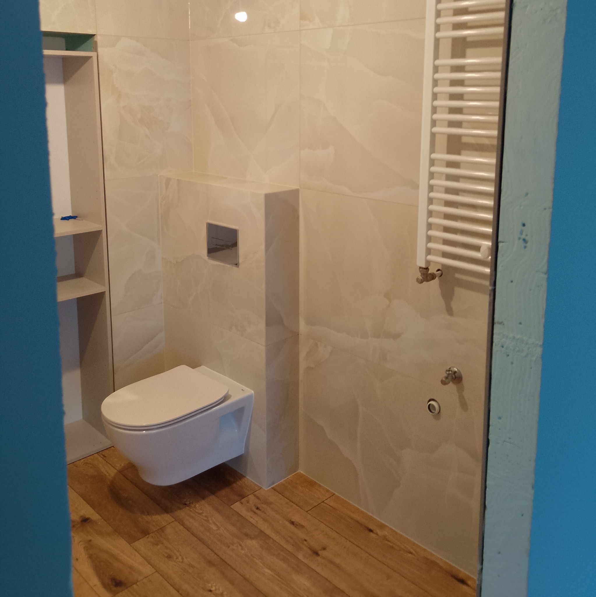 Nowoczesna łazienka z podwieszaną toaletą, grzejnikiem drabinkowym, beżowymi płytkami imitującymi marmur i drewnopodobną podłogą, widok zza niebieskiej framugi.