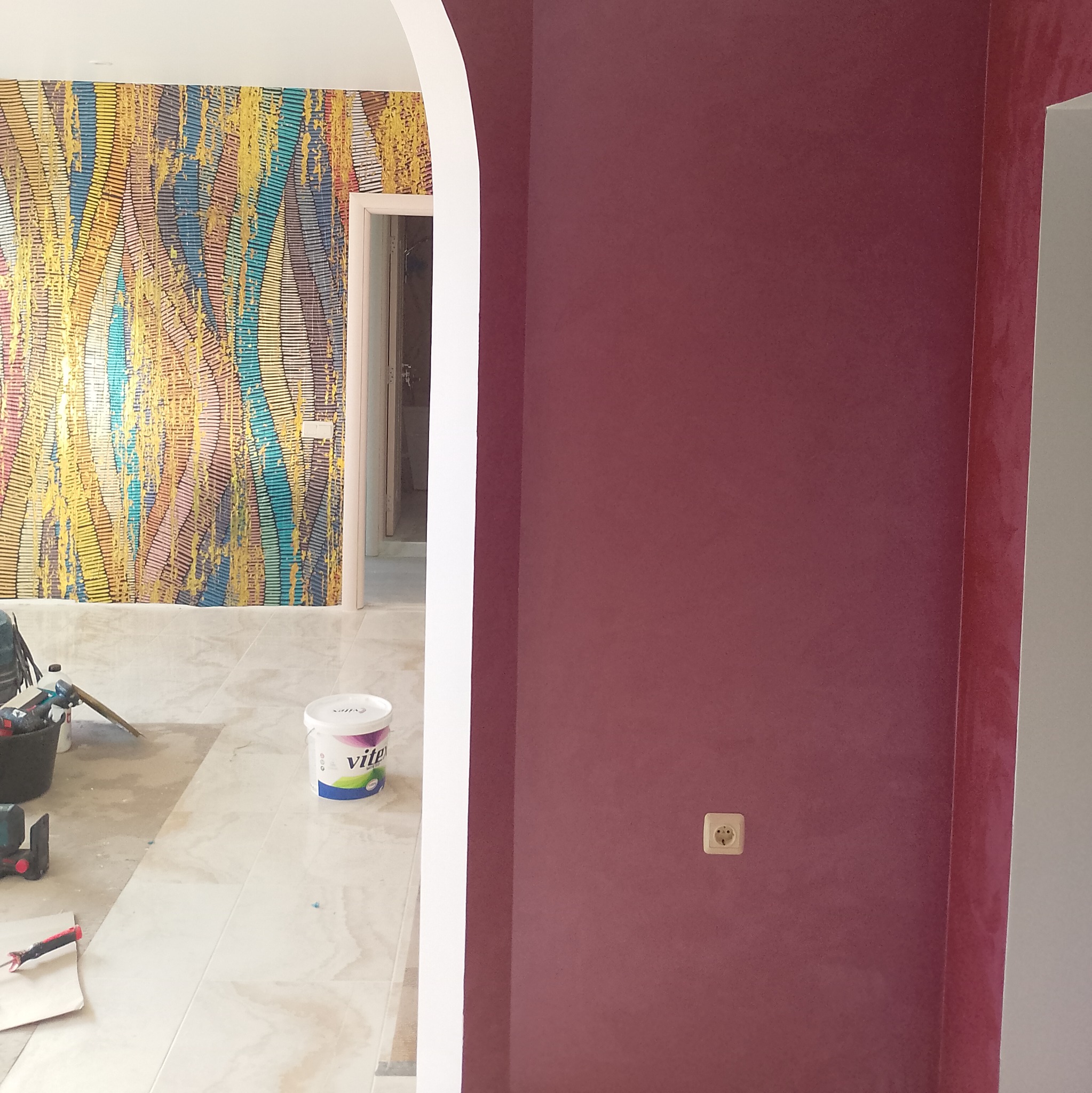 Ściana pomalowana na bordowo z widocznym gniazdkiem elektrycznym, w tle ściana z kolorową tapetą i wiadro z farbą VITE na podłodze.