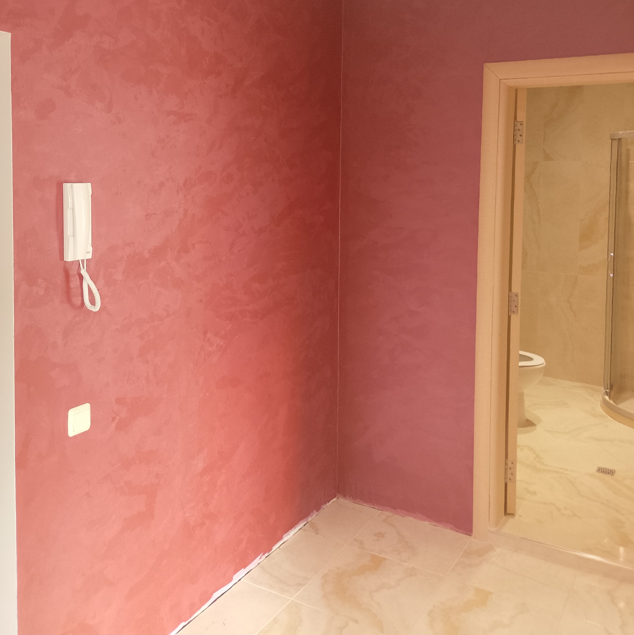 Ściana pomalowana dekoracyjną farbą w odcieniach różu, z widocznym domofonem i włącznikiem światła, oraz otwartymi drzwiami do wykończonej łazienki z kabiną prysznicową.