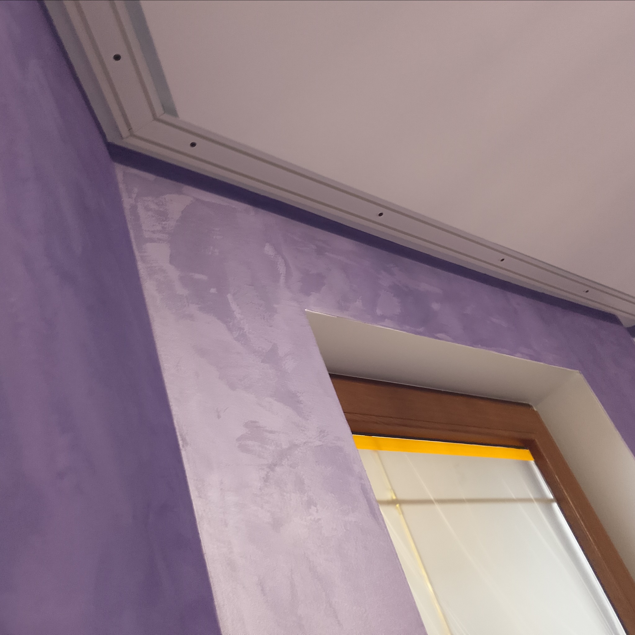 Tekstura fioletowej ściany w Kaliszu, z widoczną listwą przysufitową i fragmentem okna zabezpieczonego taśmą malarską.