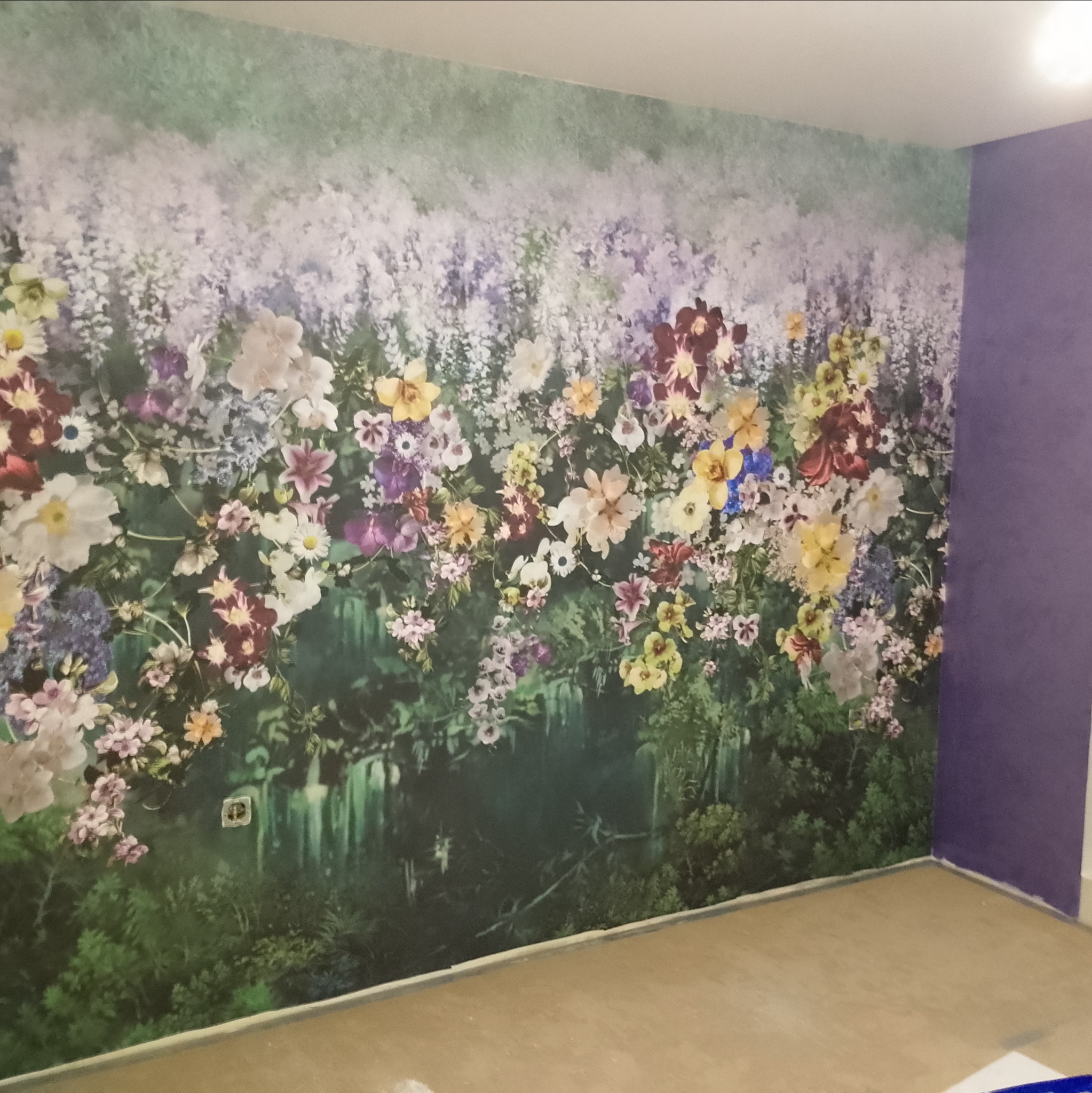 Ściana z fototapetą przedstawiającą bujny, kwiecisty motyw roślinny, sąsiadująca z fioletową ścianą, widoczny fragment podłogi w stanie surowym.