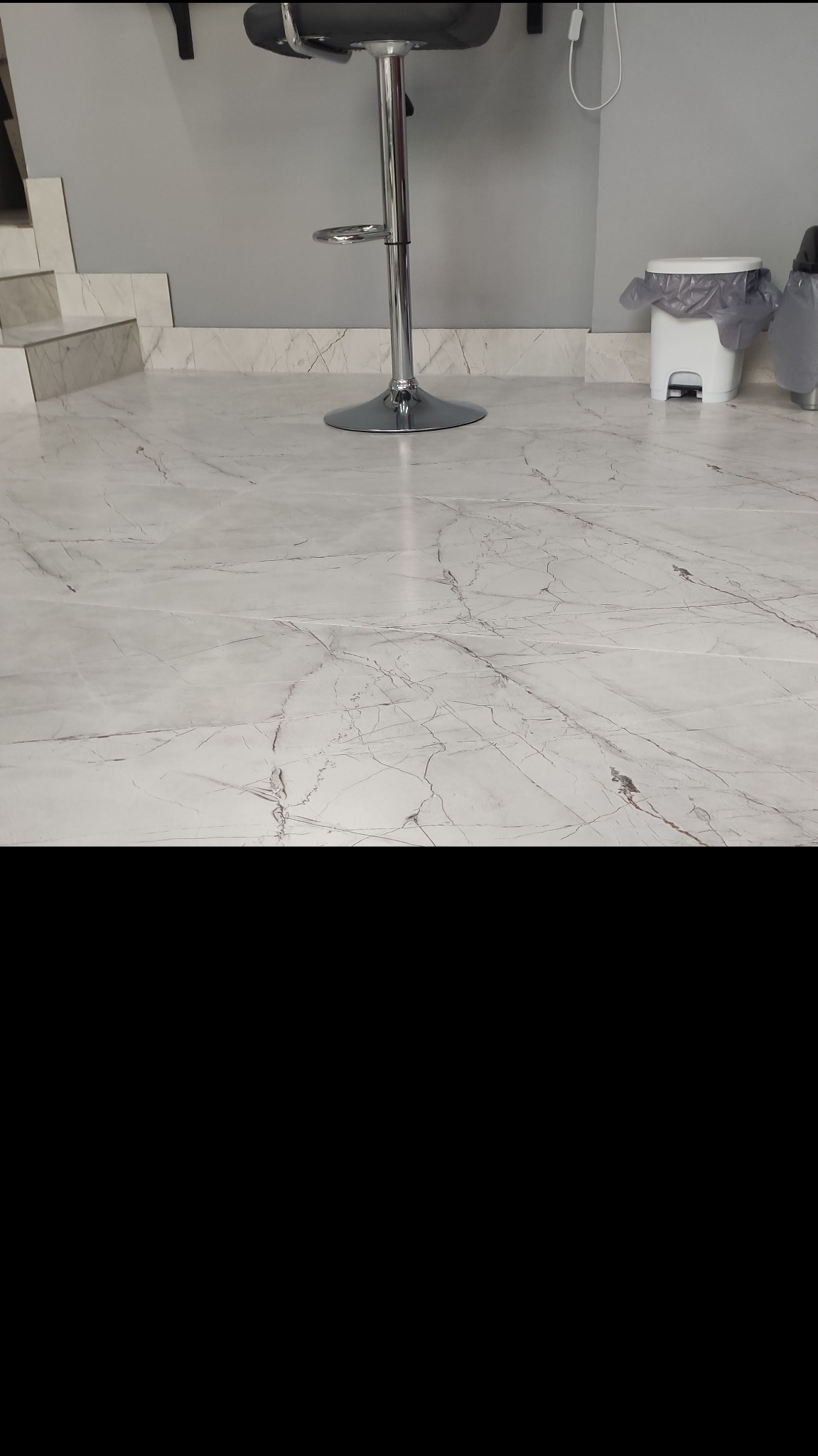 Biała podłoga z płytek imitujących marmur, widoczny fragment schodów z identycznych płytek, chromowany hoker barowy i biały kosz na śmieci w tle.