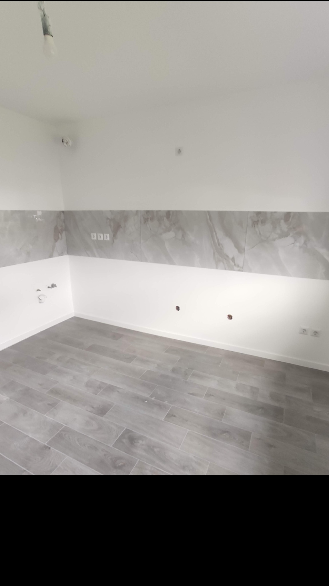 Wnętrze pokoju po częściowym remoncie z ułożonymi panelami podłogowymi w odcieniach szarości, białymi ścianami z szarym pasem płytek imitujących marmur na górze, widoczne gniazdka elektryczne...