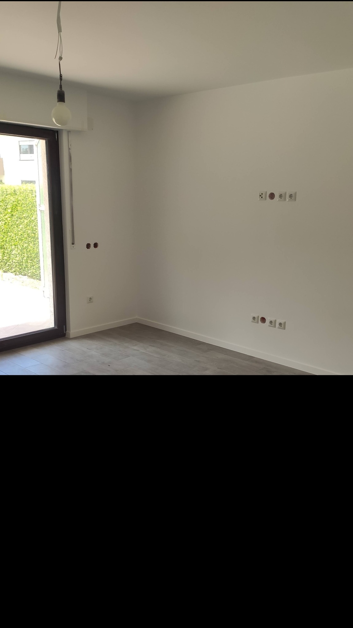 Pusty, świeżo wyremontowany pokój z szarą podłogą imitującą drewno, białymi ścianami z gniazdkami elektrycznymi i otwartym balkonem z widokiem na zielony żywopłot.