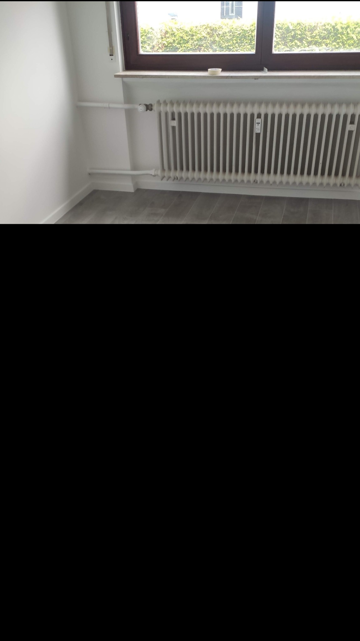 Biały, żeliwny grzejnik żeberkowy pod oknem w odnowionym pokoju, z szarymi panelami podłogowymi i białymi ścianami.