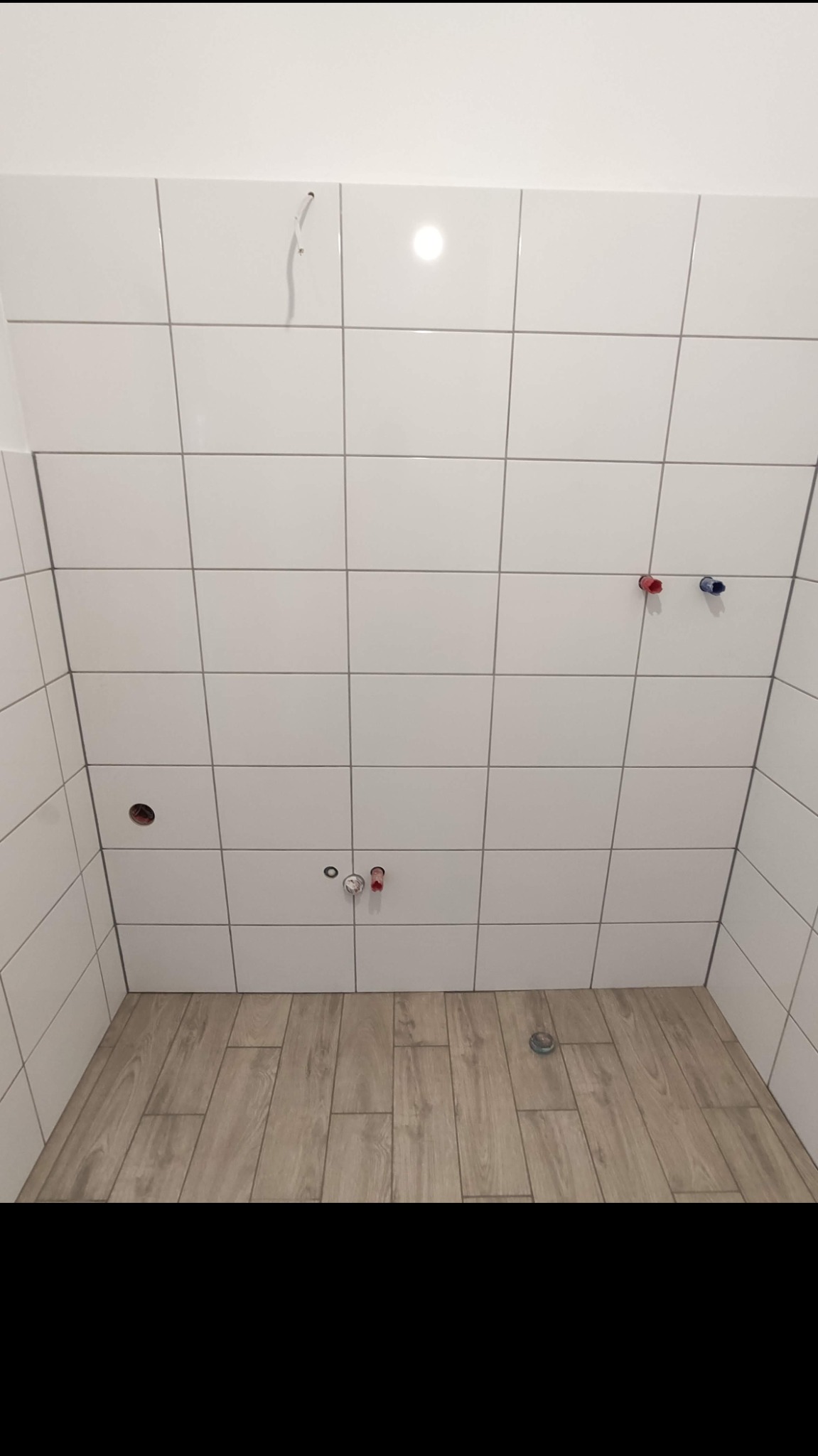 Przygotowana do wykończenia kabina prysznicowa z białymi, błyszczącymi płytkami ściennymi i podłogą wyłożoną panelami imitującymi drewno; widoczne wyprowadzenia instalacji wodnej i kanalizacyjnej.