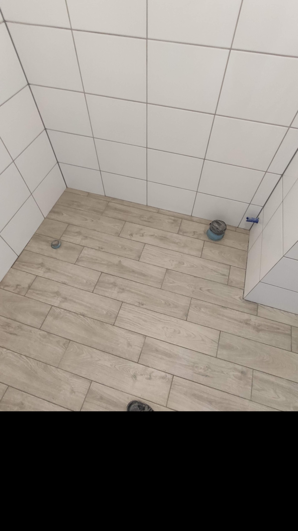 Ukończona łazienka z białymi kwadratowymi płytkami ściennymi i podłogą z paneli imitujących drewno, widoczne niebieskie zaślepki instalacyjne i odpływ podłogowy.