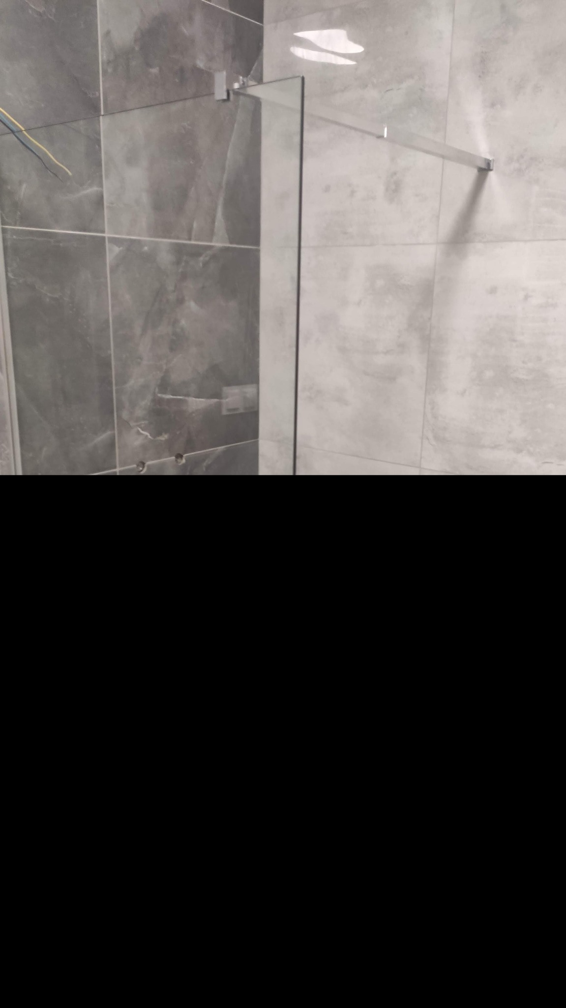 Fragment łazienki z szarymi płytkami imitującymi kamień i zamontowaną szklaną ścianką kabiny prysznicowej z chromowanymi mocowaniami.