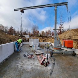 METABUDOWNICTWO SPÓŁKA Z OGRANICZONĄ ODPOWIEDZIALNOŚCIĄ - Budowa domu z bloczków: pracownik w odblaskowej kamizelce muruje ścianę fundamentową, widoczny dźwig, paleta z bloczkami, piła do cięcia bloczków oraz wózek paletowy.