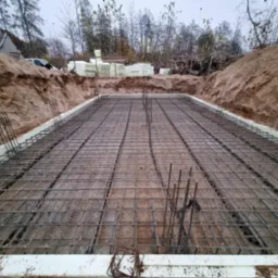 Przygotowany zbrojony szkielet fundamentu na gruncie, otoczony izolacją i wykopem ziemnym, z hałdą ziemi i stertą styropianu w tle.