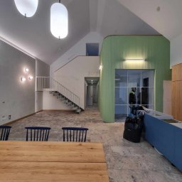 METABUDOWNICTWO SPÓŁKA Z OGRANICZONĄ ODPOWIEDZIALNOŚCIĄ - Wnętrze domu w stanie deweloperskim z drewnianym stołem na pierwszym planie, widoczne schody, surowe ściany i elementy wykończeniowe w odcieniach szarości i zieleni.