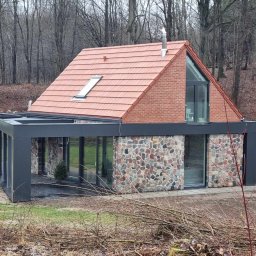 METABUDOWNICTWO SPÓŁKA Z OGRANICZONĄ ODPOWIEDZIALNOŚCIĄ - Nowoczesny dom z dachem z czerwonej dachówki, ceglaną górną kondygnacją i kamienną dolną częścią elewacji, z przeszklonymi ścianami i czarną konstrukcją tarasu, otoczony lasem i gałęziami.